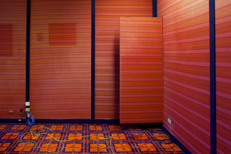 An interior corner features walls covered in horizontal orange and red striped wallpaper, accented by dark blue vertical trim and a floating panel, with a patterned orange and blue carpet and loose electrical cables on the floor.
