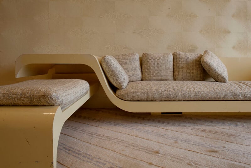 A cream-colored, curved, modern-style sofa with textured fabric cushions sits against a patterned wallpaper wall on a carpeted floor.