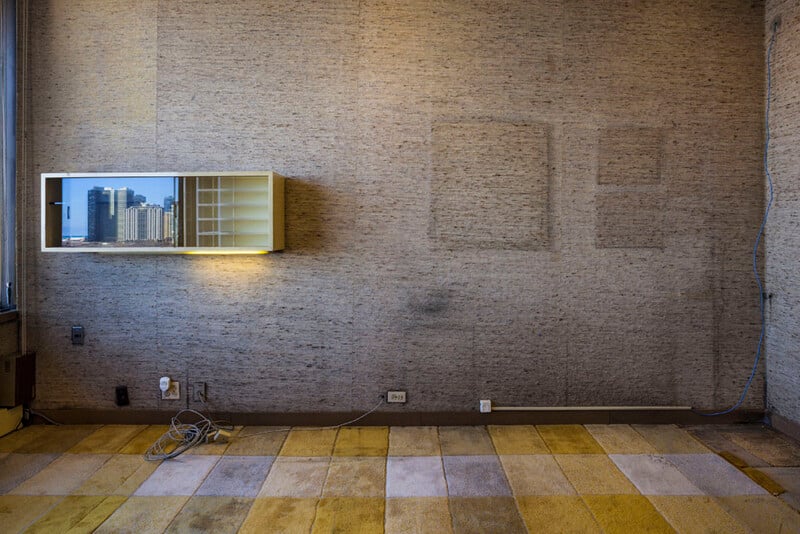 A wall with textured beige wallpaper features a rectangular, illuminated wall-mounted cabinet reflecting a city skyline, with faded rectangular outlines on the wall and a yellow and gray tiled carpet floor.