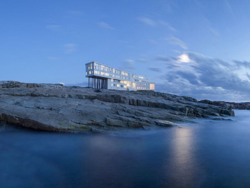 A modern, multi-story white building with many windows stands on stilts atop a rocky coastal cliff at twilight.