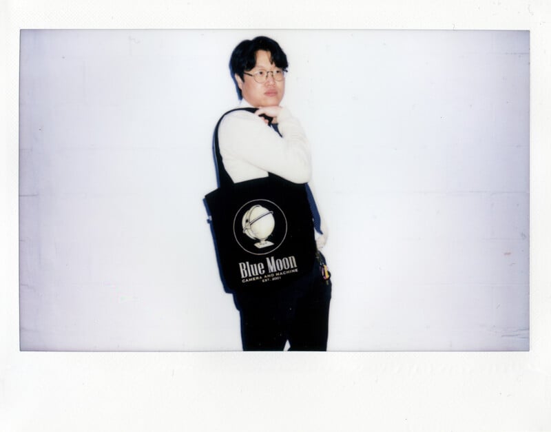 A person with dark hair and glasses wears a white sweater and black pants while carrying a black tote bag featuring a globe graphic and the text "Blue Moon Camera and Machine Est. 2001.
