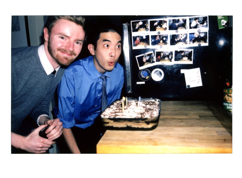 Two men stand behind a rectangular cake with lit candles on a wooden counter, with a refrigerator covered in small photos behind them.