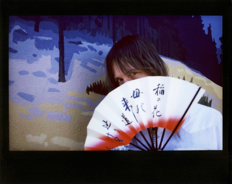 A person with shoulder-length hair holds a folding fan with Japanese calligraphy in front of their face against a painted mural of a snowy landscape.