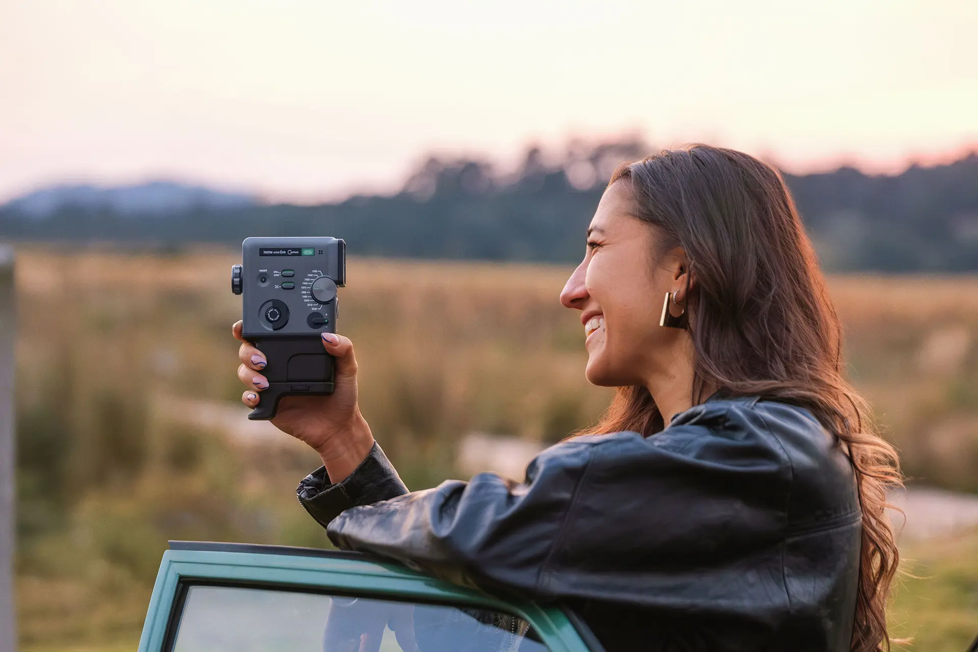 A smiling woman with long brown hair wearing a black leather jacket holds a gray digital camera while leaning against the door of a green car in a field at sunset.
