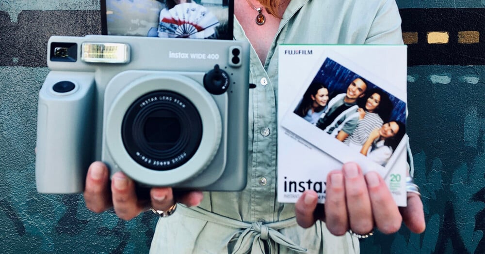 A person holds a sage green Instax Wide 400 camera in their left hand and a box of Instax instant film in their right hand.