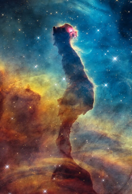 A towering, dark pillar of interstellar gas and dust rises against a vibrant, colorful nebula filled with scattered stars.