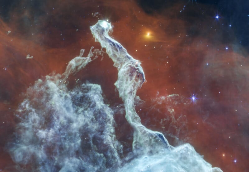 A tall, wispy pillar of glowing gas and dust rises against a backdrop of dark, reddish-orange space filled with distant stars.