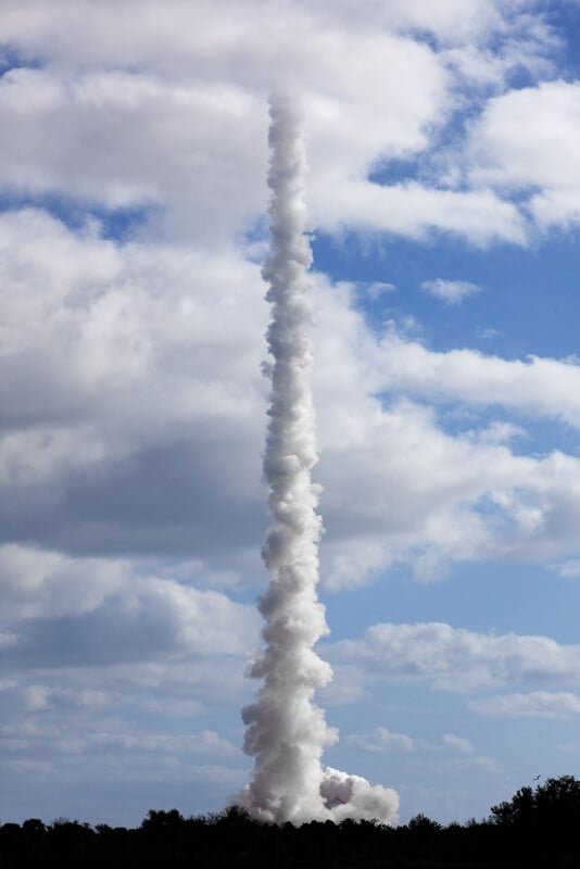 A thick, vertical column of white smoke rises from behind a dark treeline into a blue sky filled with scattered clouds.