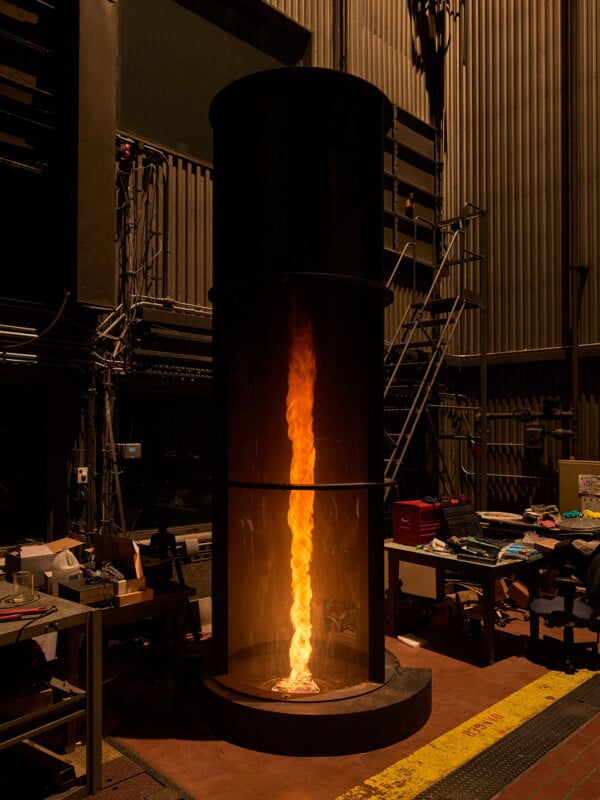 A fire tornado burns inside a cylindrical glass enclosure within a dimly lit industrial workshop.