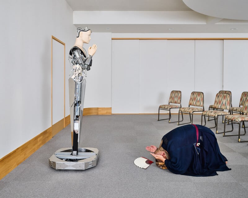 A person in traditional dark robes kneels in a deep bow before a tall, humanoid robot standing with its hands pressed together.
