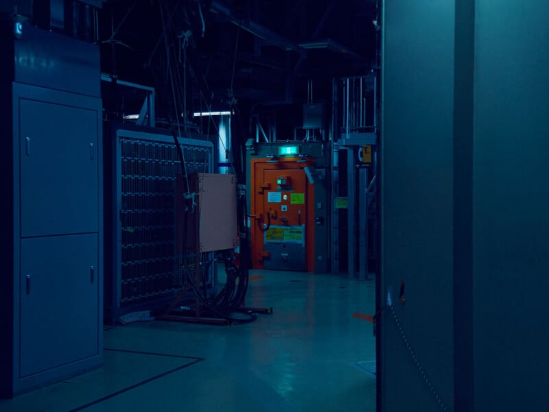 A dimly lit industrial room features a bright orange door with an illuminated green exit sign above it, surrounded by metal cabinets and electrical equipment.