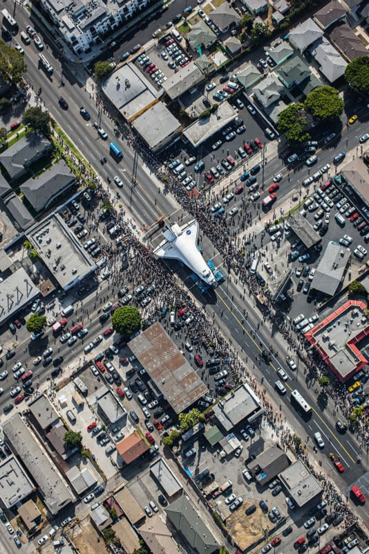 An aerial view shows a space shuttle being transported on a large trailer through a busy city intersection surrounded by crowds of people and parked cars.