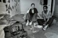 A woman and a man sit on a mattress on the floor in a room, with a trumpet case, a stack of papers, and scattered items on the carpet.