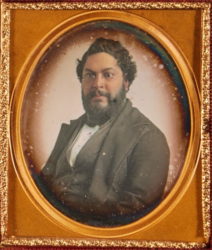 A portrait of a man with dark, curly hair and a full beard, wearing round spectacles and a dark suit, set within an ornate oval gold frame.