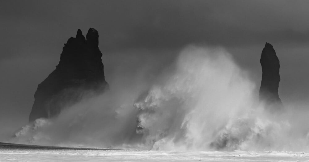 A large, powerful ocean wave crashes against dark, jagged sea stacks under a moody, overcast sky.
