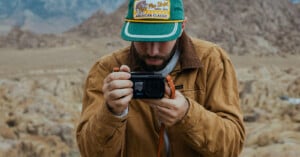 A bearded man wearing a green Bass Pro Shops cap and a brown jacket holds a small black action camera to take a photo in a rocky, desert landscape.