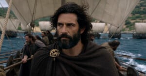A bearded man with dark, wavy hair wears a brown cloak fastened with a gold brooch while standing on a ship at sea.