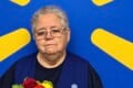 An older woman with short gray hair and glasses wears a navy blue vest over a dark shirt, holding a bouquet of red and yellow flowers in front of a blue wall with a yellow sunburst pattern.