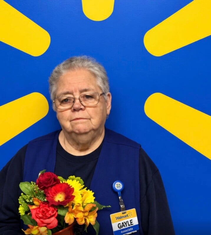 An older woman with short gray hair and glasses wears a navy blue vest over a dark shirt, holding a bouquet of red and yellow flowers in front of a blue wall with a yellow sunburst pattern.
