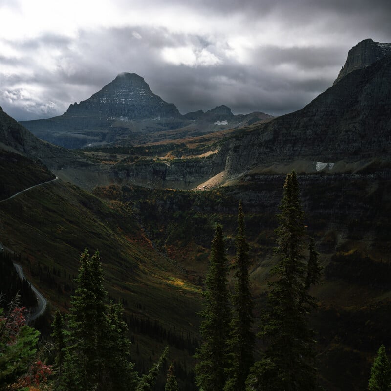 A winding road cuts through a deep mountain valley under a dramatic, cloudy sky, with evergreen trees in the foreground.