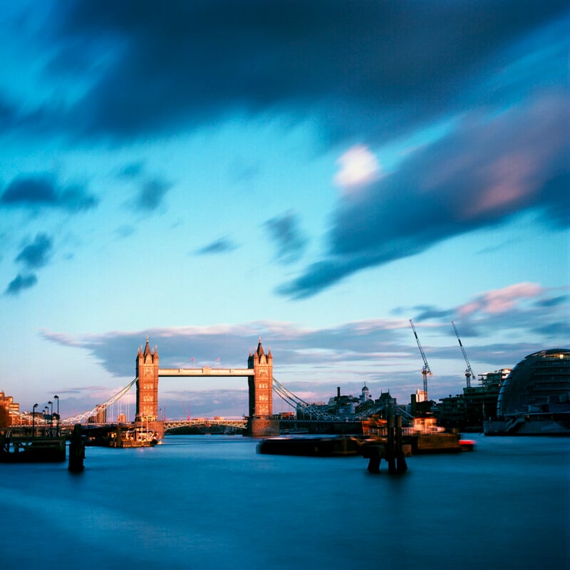Tower Bridge stands over the River Thames at twilight under a cloudy blue sky, with construction cranes visible nearby.