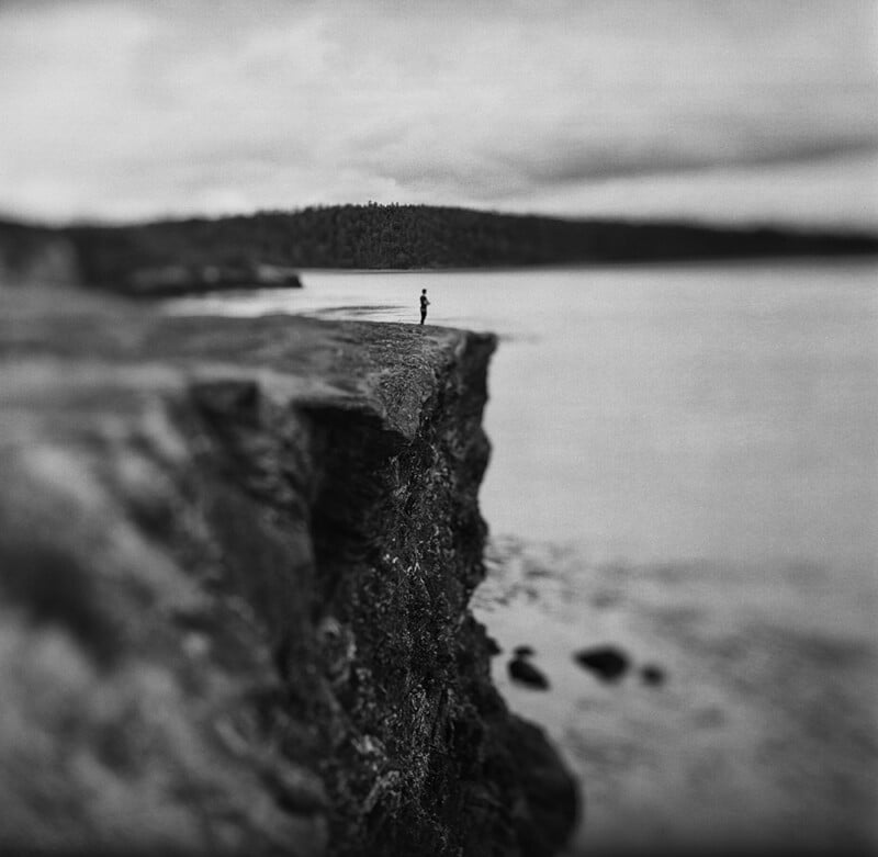 A lone figure stands on the edge of a rocky cliff overlooking a calm body of water under a cloudy sky.