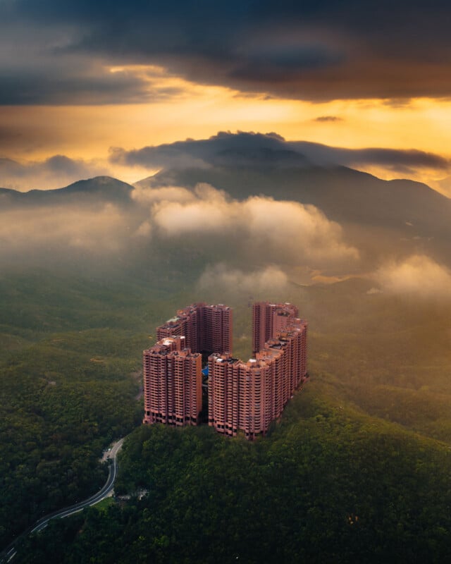 An aerial view of a large, U-shaped pink apartment complex nestled in a lush, green mountain range under a dramatic, cloudy sunset sky.