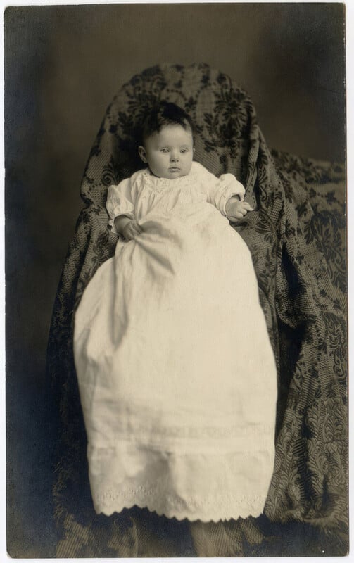 A baby in a long white gown sits upright against a dark, patterned fabric backdrop.