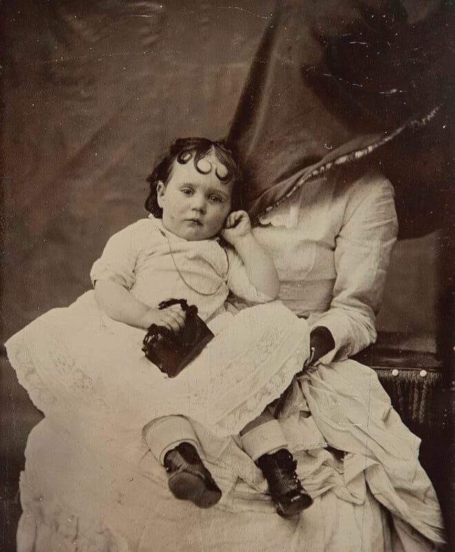 hidden mother style portrait of a young child and her mother, whose face is obscured by a cloth