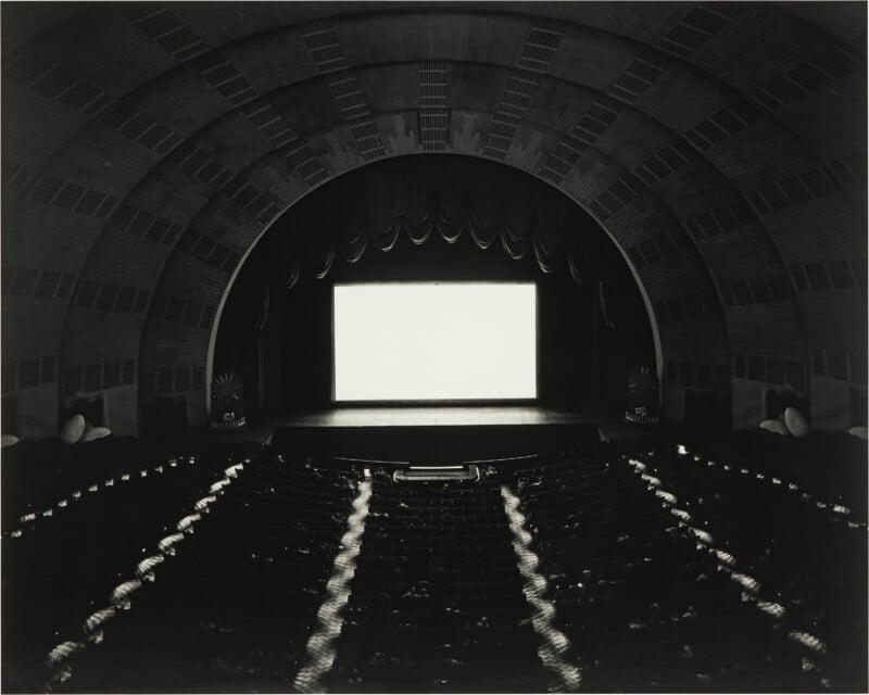 A wide-angle, black-and-white view of a large, arched theater interior with a brightly lit, blank movie screen.
