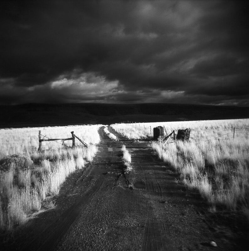 A dirt road leads through a grassy field toward a dark, cloudy sky, framed by a wire fence and wooden gate posts.