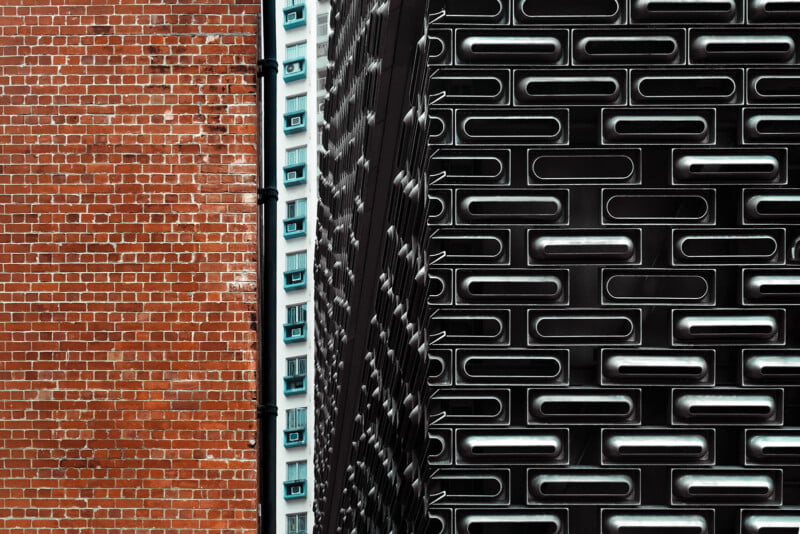 A red brick wall stands next to a white building with blue window frames and a modern, dark gray facade with repeating slots.