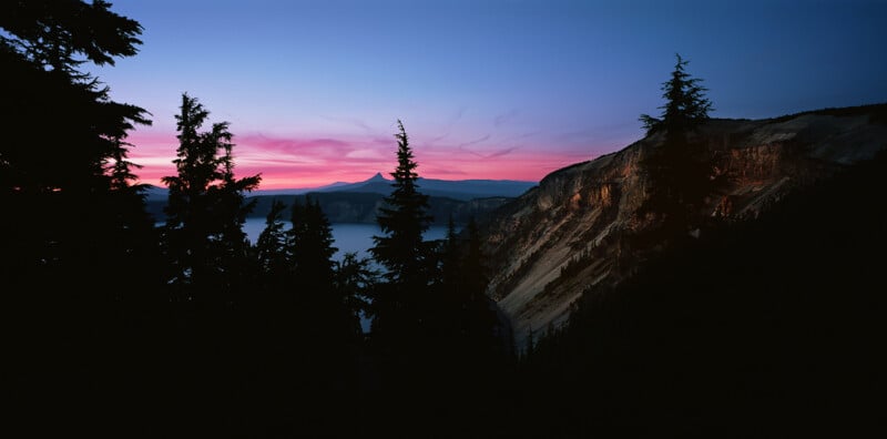 Silhouetted evergreen trees frame a view of a lake and distant mountain peak under a vibrant pink and blue sunset sky.