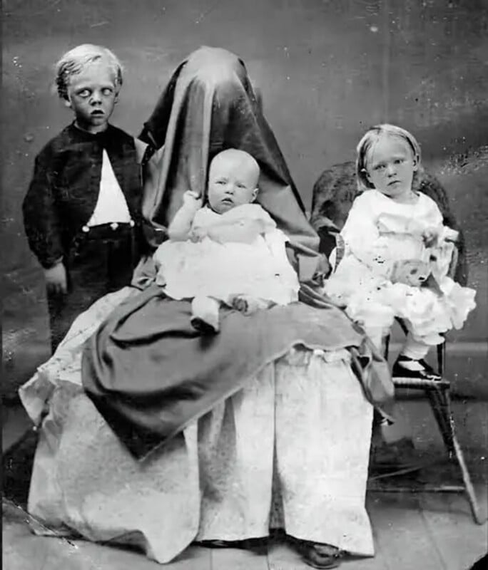 A vintage black-and-white portrait of a mother covered by a dark cloth, holding a baby, flanked by a young boy and girl.