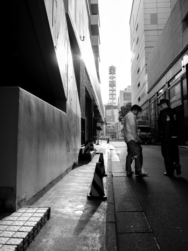 Two men stand on a narrow city street between tall buildings, with traffic cones lining the sidewalk and a communication tower visible in the distance.