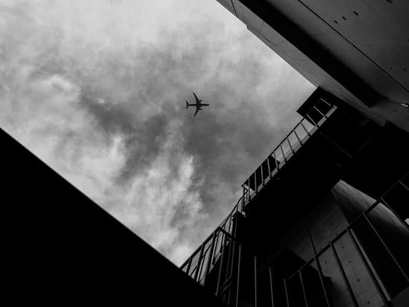A low-angle, black and white shot looking up at an airplane flying through a cloudy sky, framed by the corner of a building.