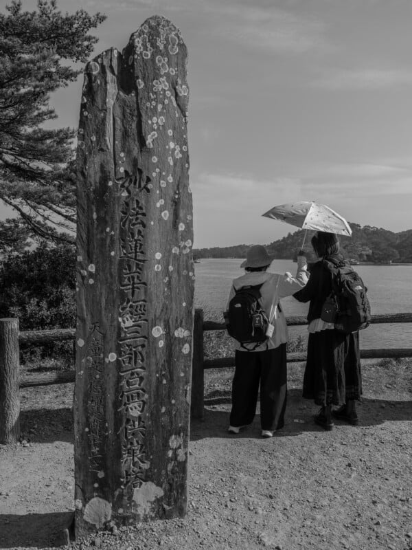 Two people stand by a wooden fence overlooking a body of water, with one holding a parasol and a tall, weathered stone monument inscribed with Japanese characters in the foreground.