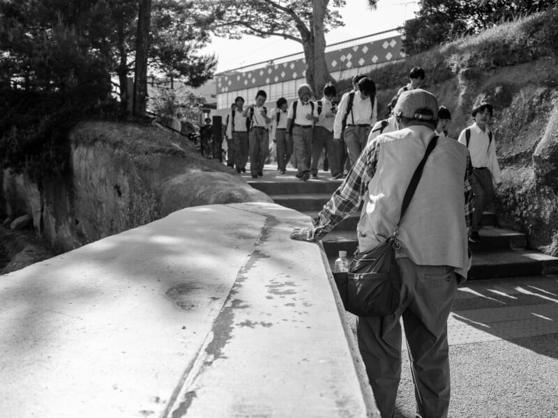 An older person with a shoulder bag stands by a stone wall as a group of schoolboys in uniforms walks up a path behind them.