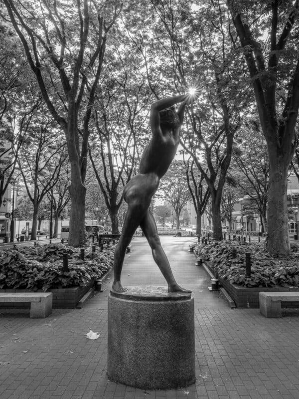 A bronze statue of a nude figure stands on a pedestal in a tree-lined park, with the sun shining directly above its raised hands.