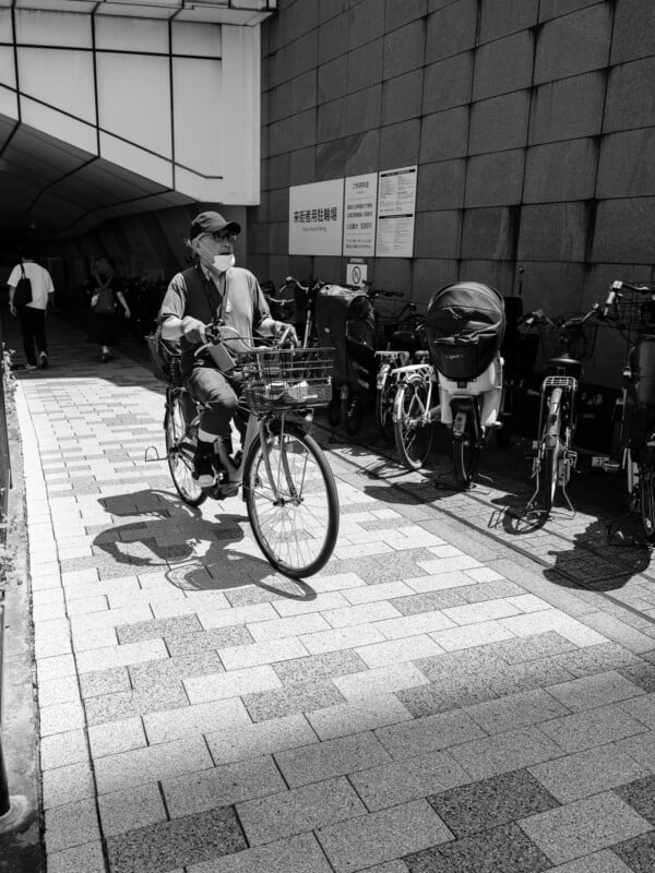 An older man wearing a cap and face mask rides a bicycle past a row of parked bikes and a wall with informational signs.
