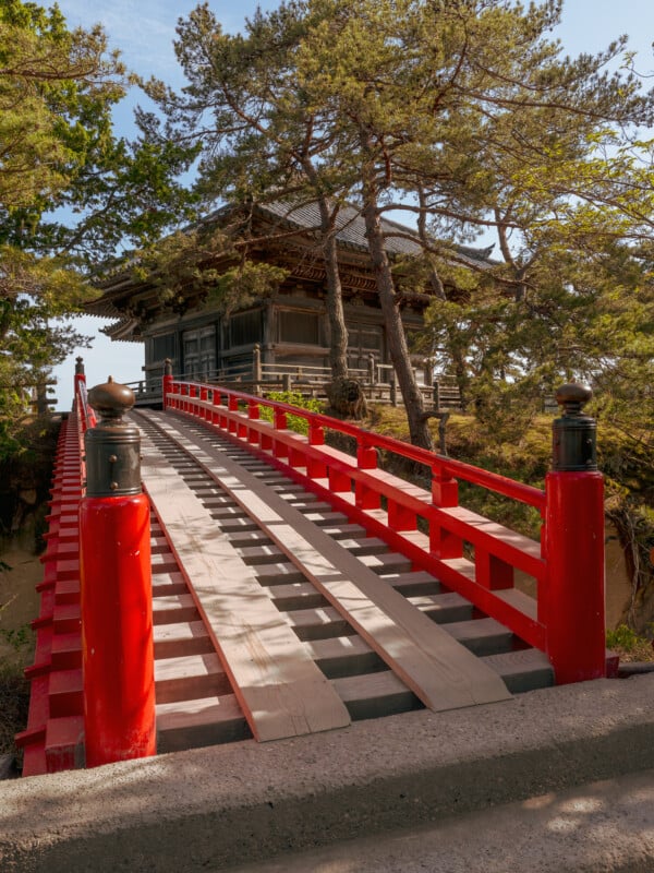 A red arched bridge with wooden planks leads toward a traditional wooden temple building surrounded by pine trees.