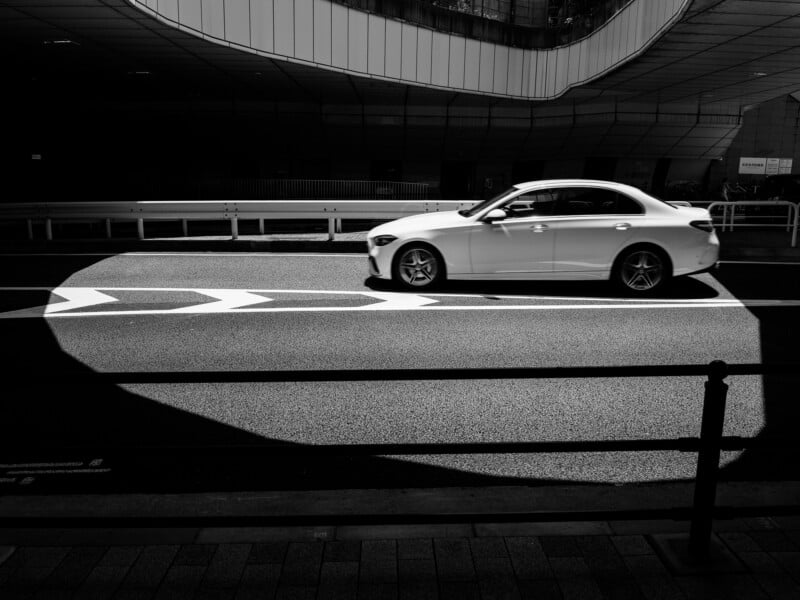 A white sedan drives across a sunlit patch of asphalt, framed by deep shadows and a metal railing in the foreground.
