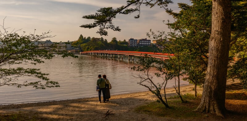 Two people stand on a sandy beach looking out at a long, red bridge spanning a calm body of water.