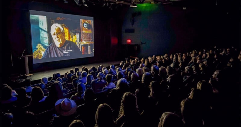 An audience sits in a dark theater watching a large screen showing a man named Herb Ascherman.