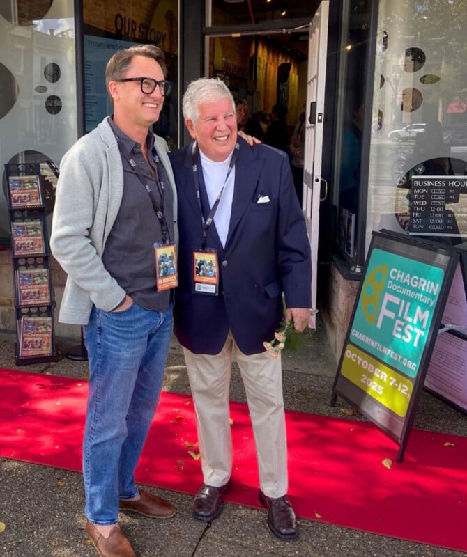 Two men stand on a red carpet in front of a building with a sign for the Chagrin Documentary Film Festival, October 7-12, 2025.