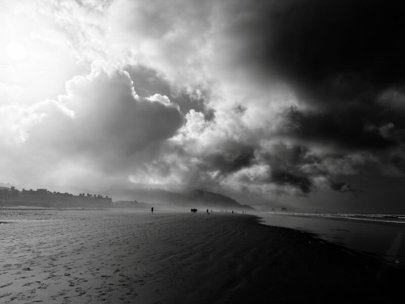 A black and white view of a wide, sandy beach under a dramatic, cloudy sky with a group of people walking in the distance.
