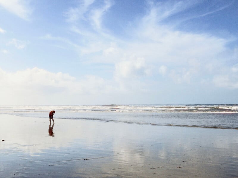 A person in a red shirt stands on a wet, reflective beach looking down at the water as waves roll in under a cloudy sky.