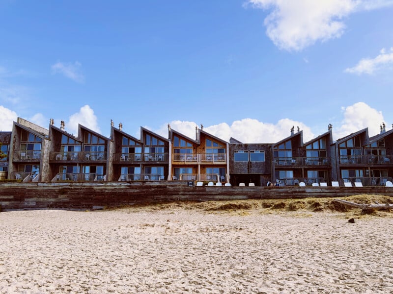 A row of multi-story, shingled beach houses with slanted roofs sits behind a wooden retaining wall above a sandy beach under a blue sky with scattered clouds.
