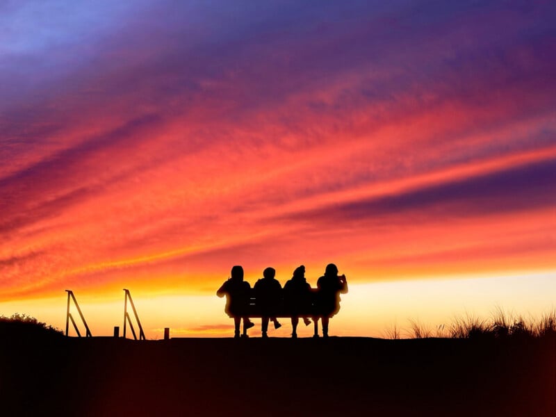 Four people sit on a bench in silhouette against a vibrant, multi-colored sunset sky.
