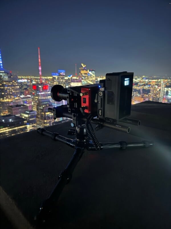 A professional cinema camera mounted on a tripod overlooks a blurred, illuminated city skyline at night.